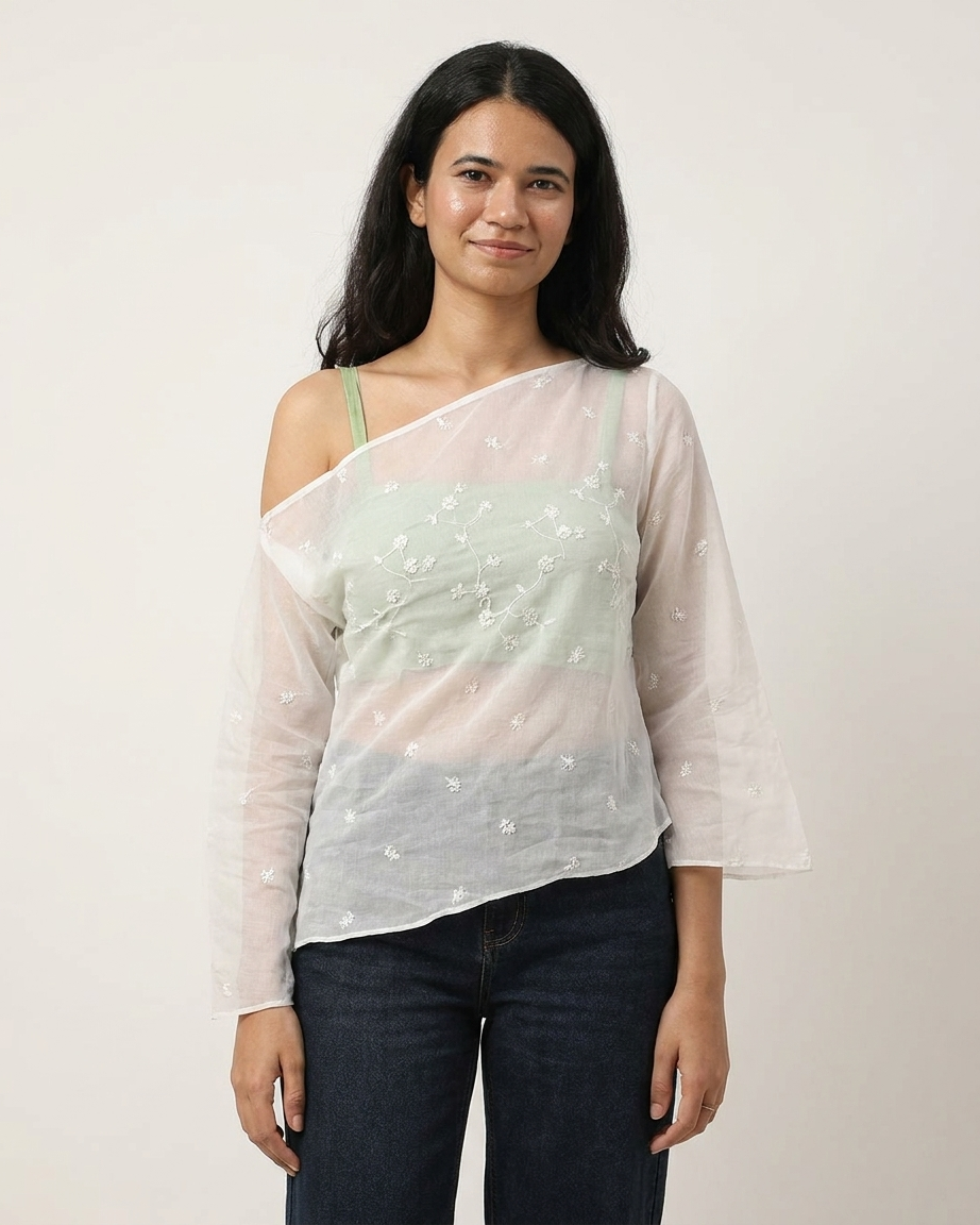 A woman modeling a sheer, asymmetrical off-the-shoulder top in white with delicate floral embroidery, worn over a pale green camisole and paired with dark blue jeans, against a plain light background.