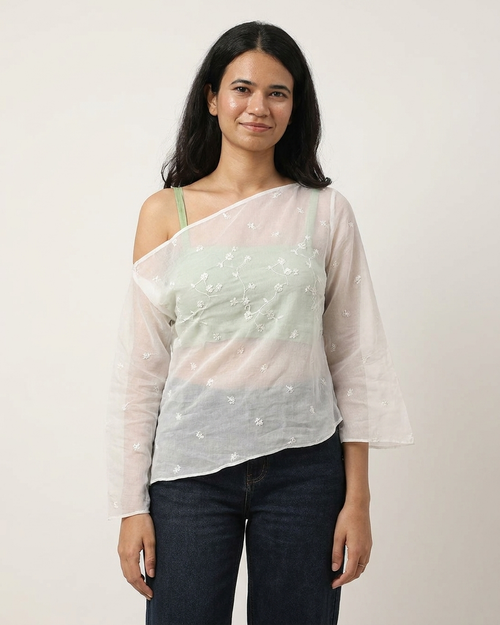 A woman modeling a sheer, asymmetrical off-the-shoulder top in white with delicate floral embroidery, worn over a pale green camisole and paired with dark blue jeans, against a plain light background.